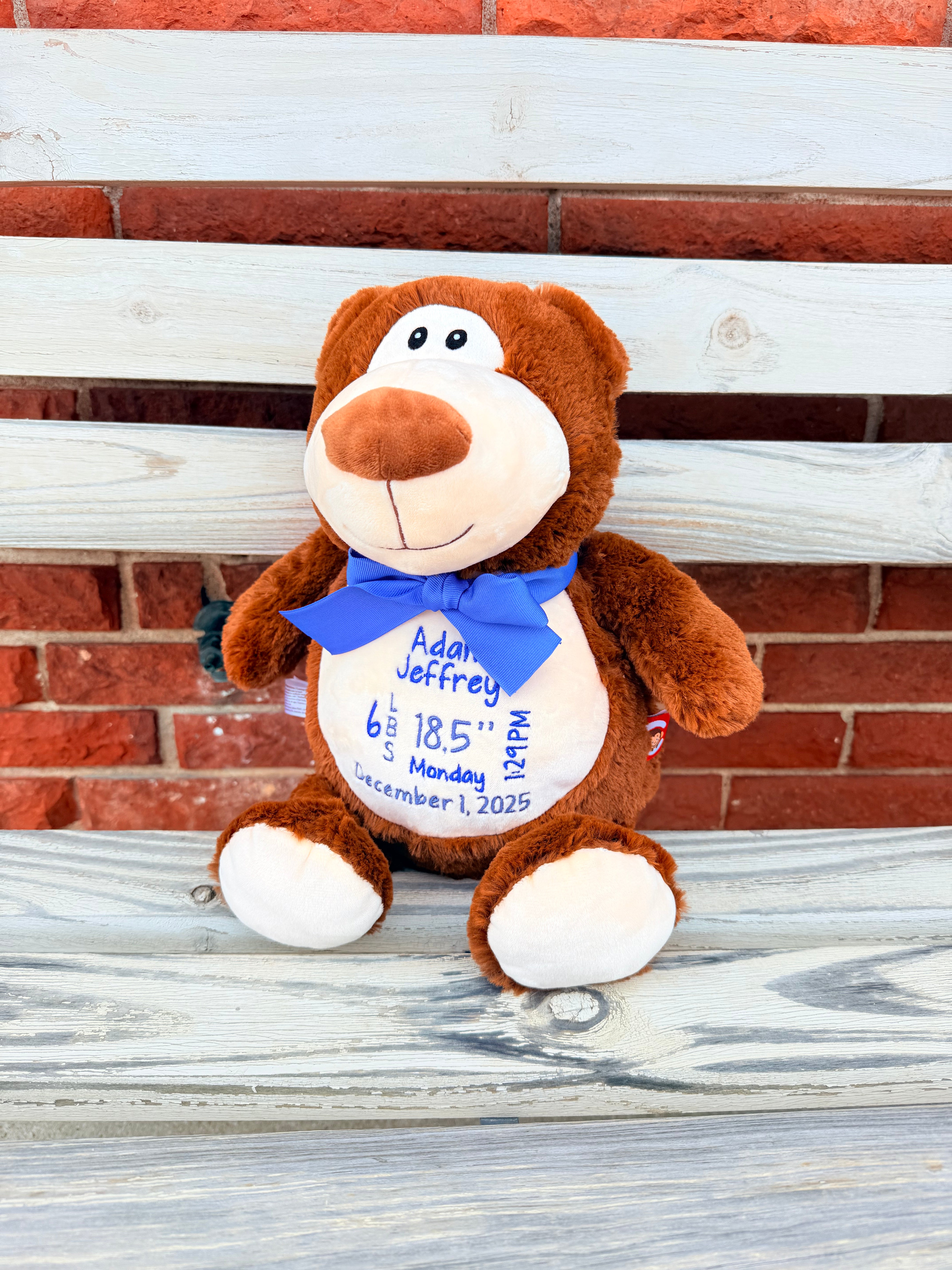 Brown teddy bear with a blue bow tie sitting on a wooden surface with a brick wall background.