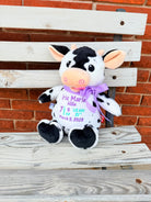 Plush cow with a bow on a wooden bench against a brick wall that is embroiered with babys birth info.