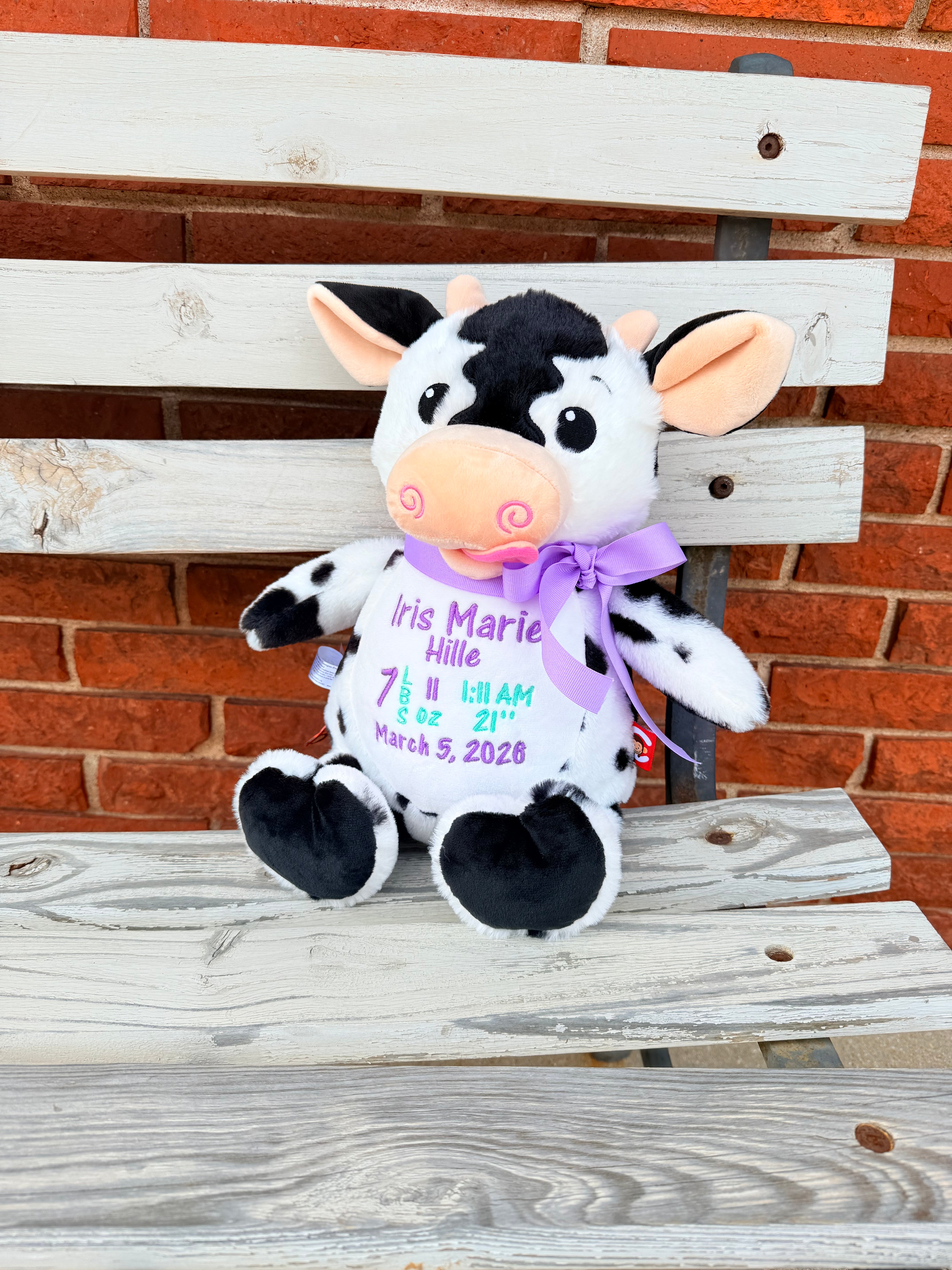 Plush cow with a bow on a wooden bench against a brick wall that is embroiered with babys birth info.