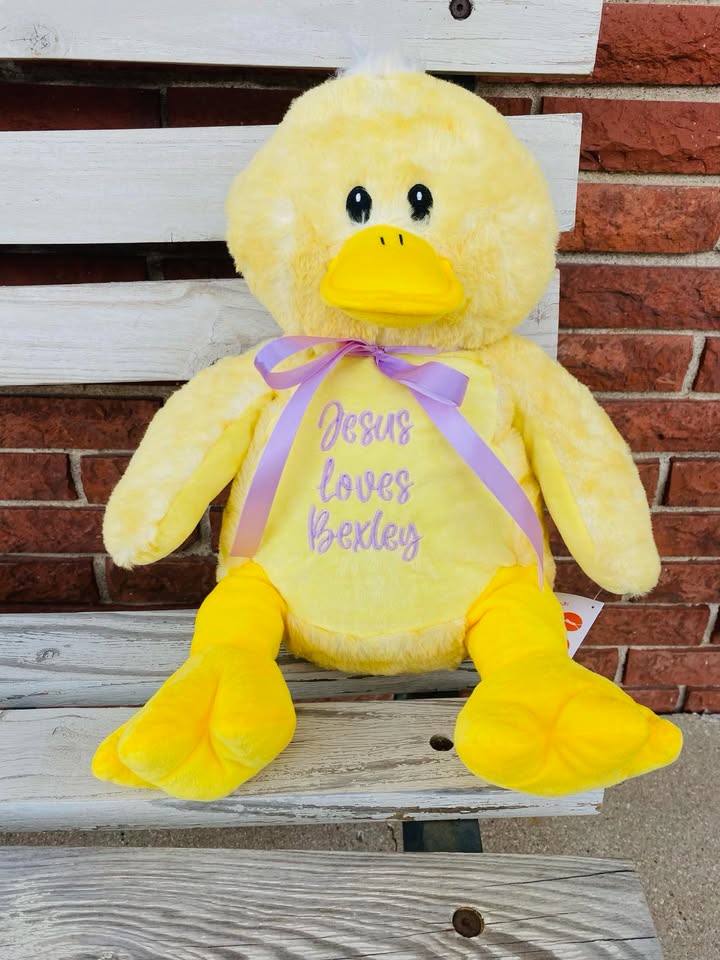 Yellow plush duck with 'Jesus loves Lexley' text on a wooden surface with brick wall background