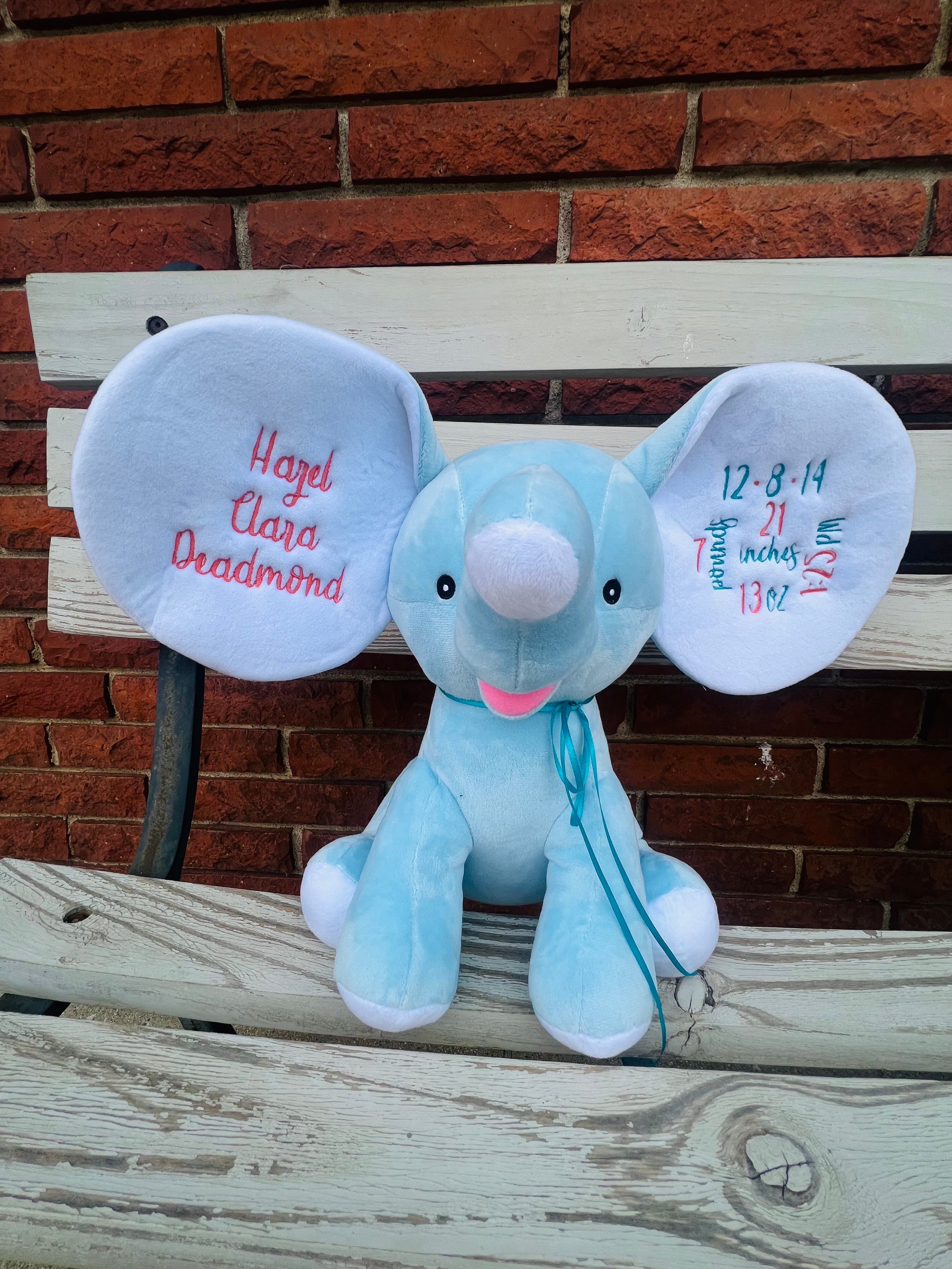 Plush elephant toy with embroidered text on ears against a brick wall.