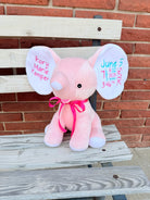Pink elephant plush toy with personalized text on ears, sitting on a wooden bench against a brick wall.