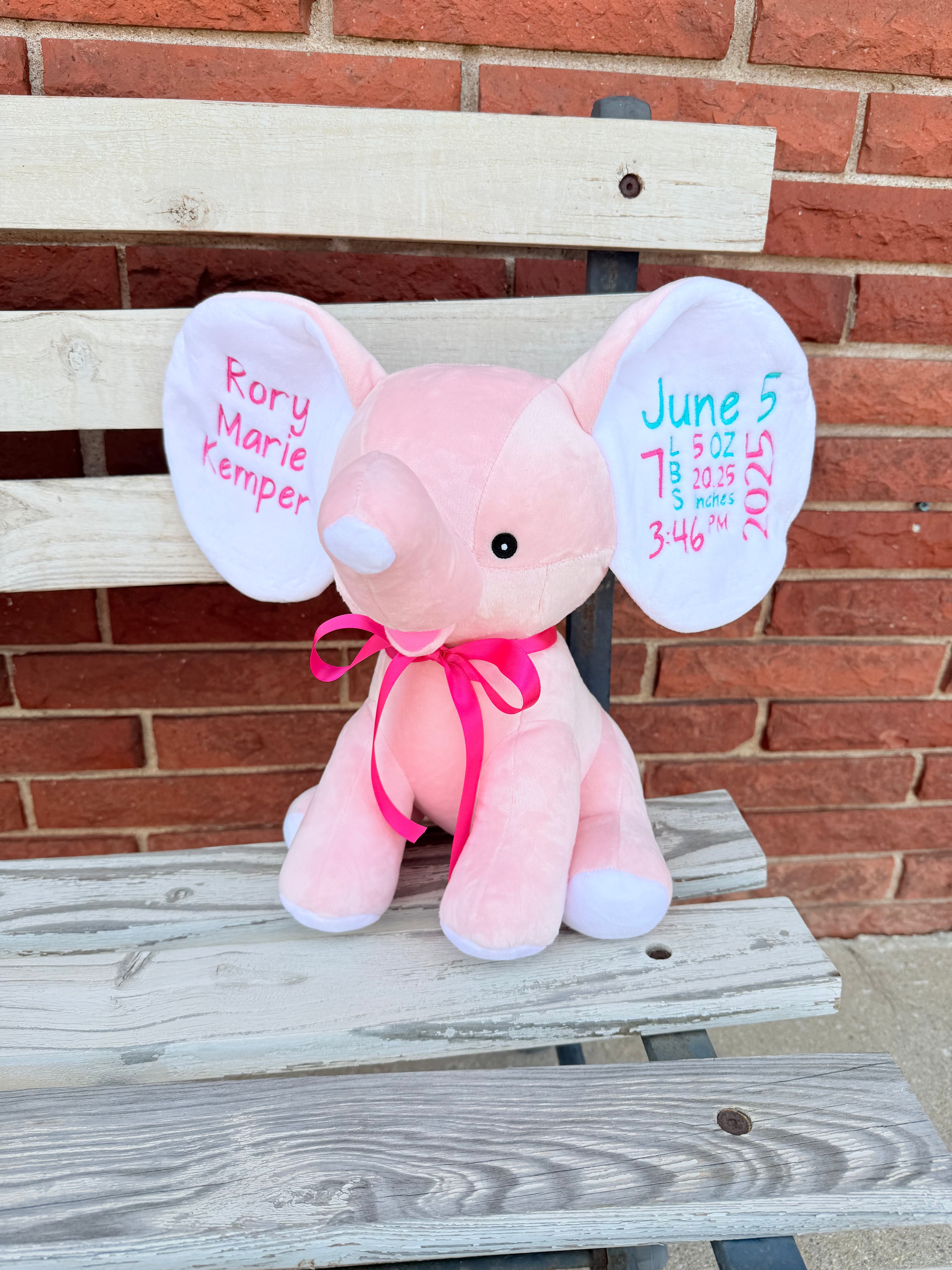 Pink elephant plush toy with personalized text on ears, sitting on a wooden bench against a brick wall.