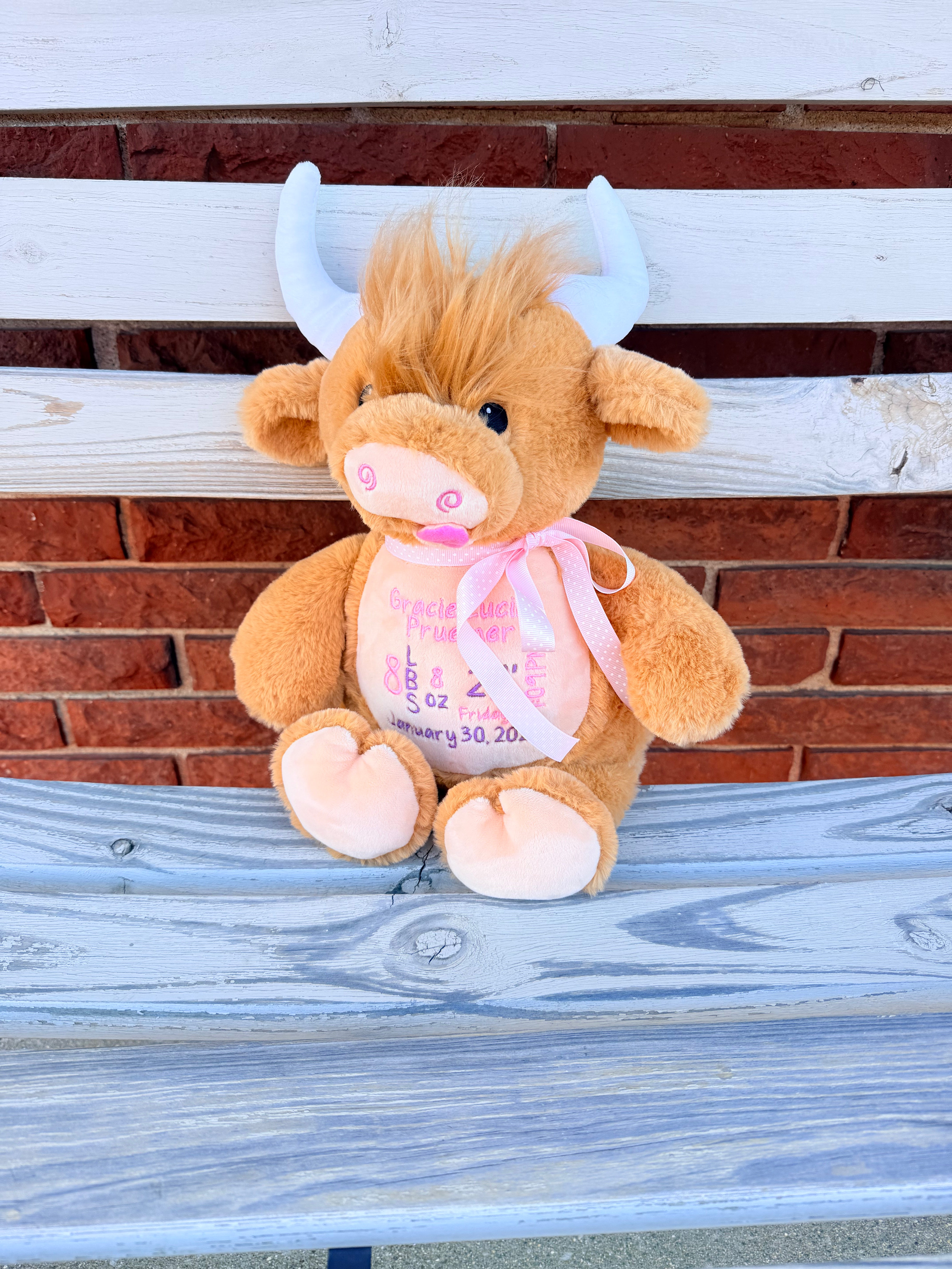 Plush cow toy with a pink bow sitting on a wooden bench against a brick wall.